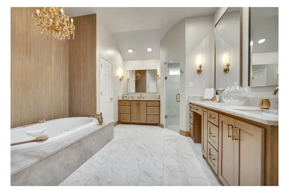 Bathroom remodeling Midland TX: Highland Residence primary bath with a drop-in soaking tub, wood vanity cabinets, marble-look floor and brass sconces