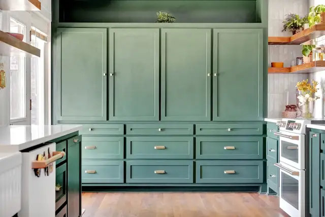 Green shaker cabinets with brass pulls, white double wall ovens and floating wood shelves in the Wydewood Residence kitchen